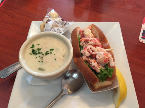 A plate with a lobster roll on a bun, lettuce, and a bowl of clam chowder, garnished with a lemon wedge.