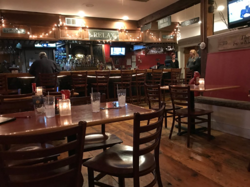 A cozy restaurant interior with wooden tables, chairs, and a bar area, softly lit with candles and string lights.