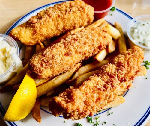 A plate of golden-brown fried fish fillets on a bed of French fries, served with lemon and dipping sauces.