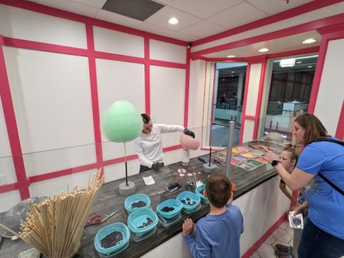 A child watches as a staff member prepares cotton candy at a colorful dessert counter, with various treats displayed.