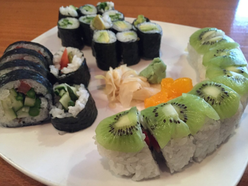A plate of sushi rolls, including cucumber and tomato, topped with kiwi slices, accompanied by wasabi and pickled ginger.