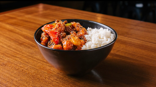 A black bowl filled with colorful stir-fried vegetables and meat, served with a side of white rice on a wooden table.