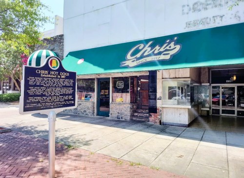 Exterior of Chris's Hot Dogs, featuring a green awning and a historical sign, located on a sunny street.