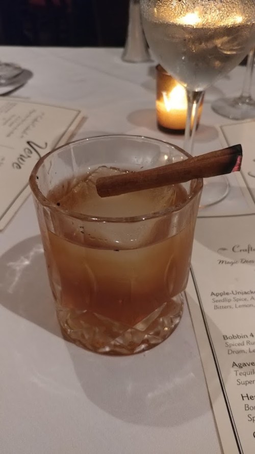 A cocktail in a crystal glass with a cinnamon stick, sitting on a table with menus and a candle in the background.