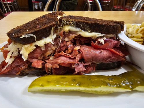 A hearty sandwich with layers of corned beef and sauerkraut on dark rye bread, served with a pickle on the side.