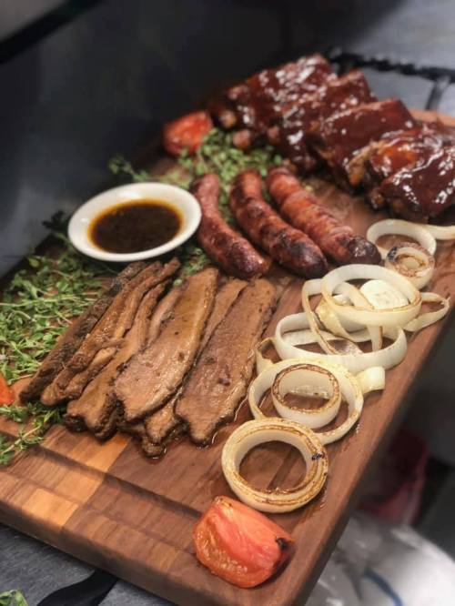 A wooden platter featuring grilled meats, including ribs, sausage, and sliced onions, garnished with herbs and sauce.