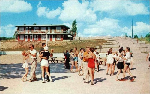 A vintage scene of couples dancing outdoors near a building, with a sunny sky and picnic tables in the background.