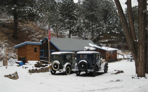 Two vintage cars parked in front of a rustic cabin surrounded by snow-covered trees. An American flag is visible.