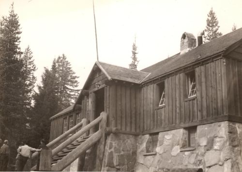 Historic wooden building with stone foundation, surrounded by trees, featuring a staircase and broken windows.
