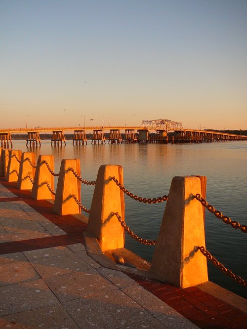 A serene waterfront scene at sunset, featuring a pier and a chain-link fence along the calm water.