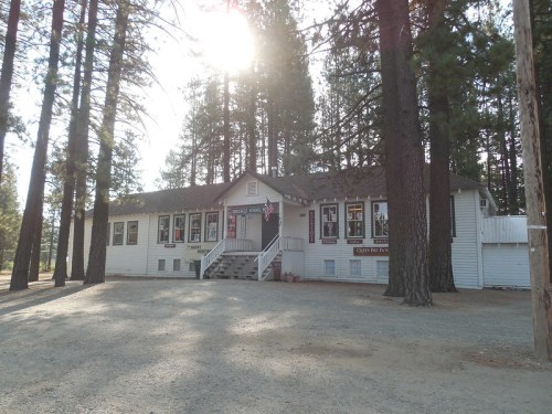 A white building with a porch, surrounded by tall trees, in a gravel area, likely a school or community center.