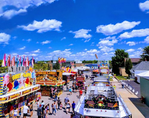 A vibrant fair scene with colorful booths, food stalls, and a crowd under a bright blue sky with fluffy clouds.