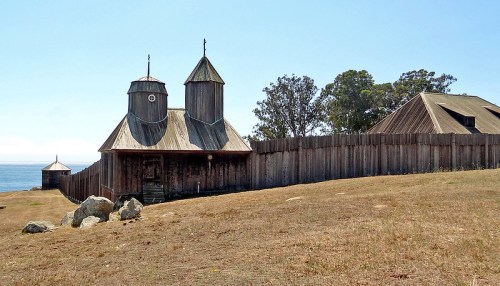 A wooden building with two towers, surrounded by a fence, near the ocean on a sunny day.