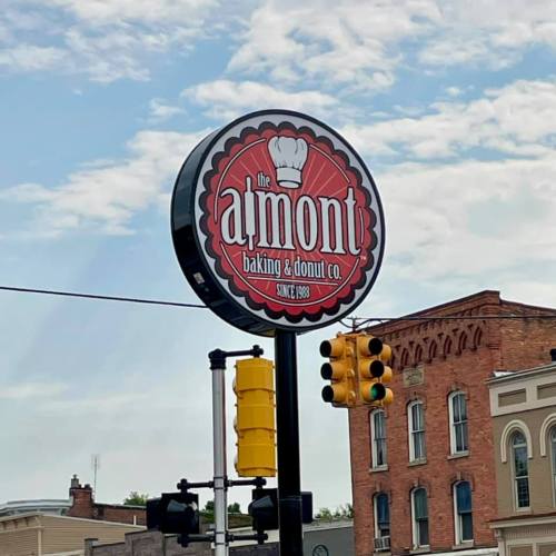 Sign for "The Almont Baking & Donut Co." with a chef's hat logo, established in 1998, against a cloudy sky.