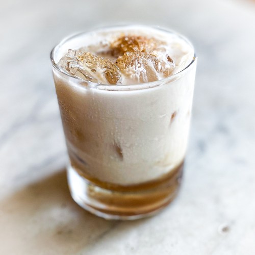 A glass of creamy iced drink with ice cubes and a sprinkle of brown sugar on top, set on a marble surface.