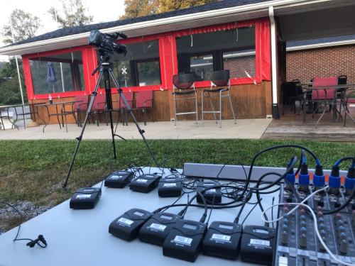 A camera on a tripod in front of a building with red curtains, alongside a table with audio equipment and multiple devices.