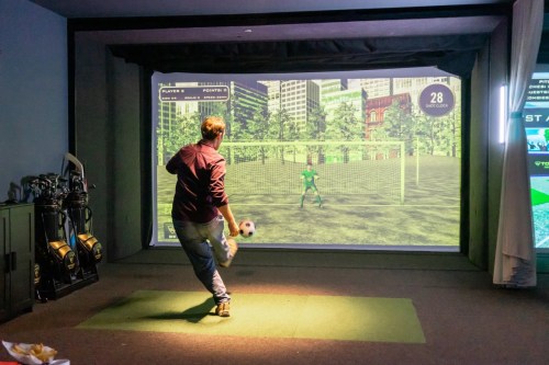 A person kicks a soccer ball towards a virtual goal on a large screen in a sports simulation room.