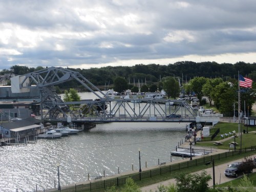 A drawbridge over a river with boats docked nearby, surrounded by trees and a cloudy sky. An American flag is visible.