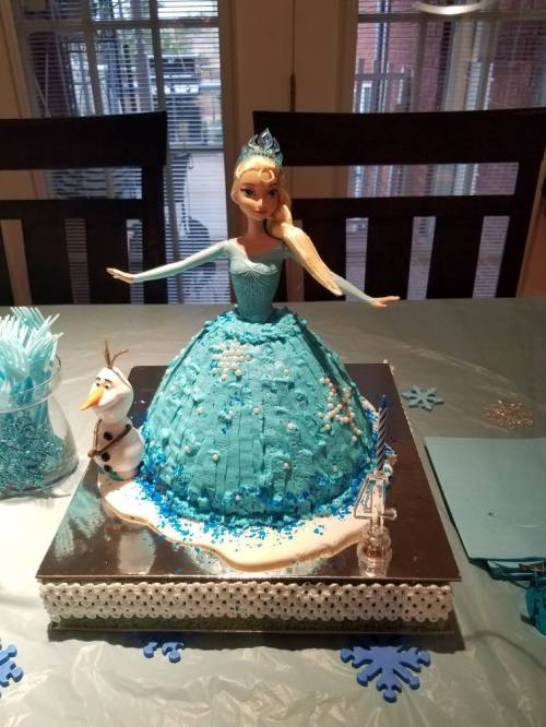 A cake shaped like a princess in a blue dress, with a snowman figure beside it, set on a decorated table.