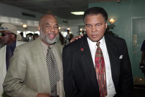 Two men in formal attire pose together in a crowded indoor setting. One man has a serious expression.