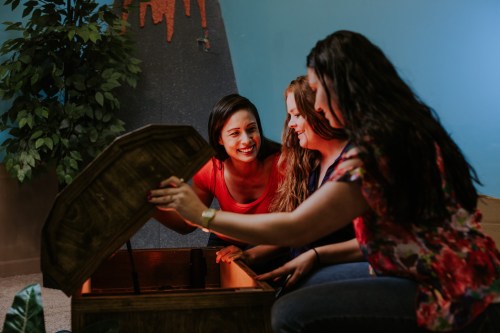 Three women excitedly look into an open treasure chest, surrounded by a colorful backdrop and indoor plants.