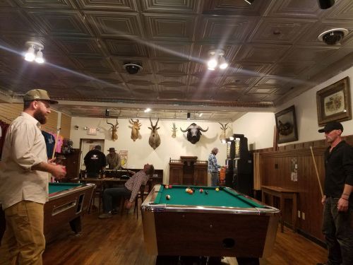 Two men play pool in a rustic bar with mounted animal heads on the walls and patrons in the background.