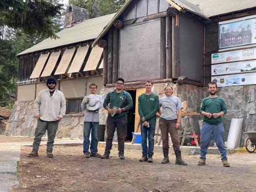 A group of six people stands in front of a rustic building, smiling and posing for the photo.