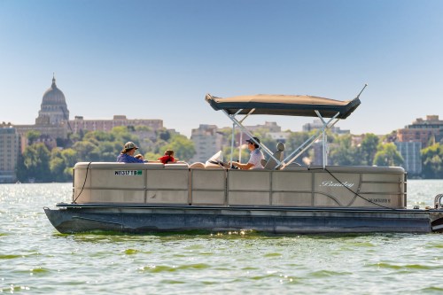 A pontoon boat with people enjoying a sunny day on the water, with a city skyline in the background.