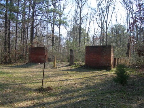 Abandoned brick structures in a wooded area, surrounded by trees and overgrown grass.