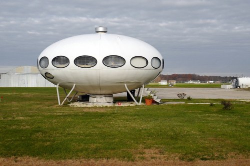 A futuristic, oval-shaped white building with large circular windows, set on a grassy area under a cloudy sky.