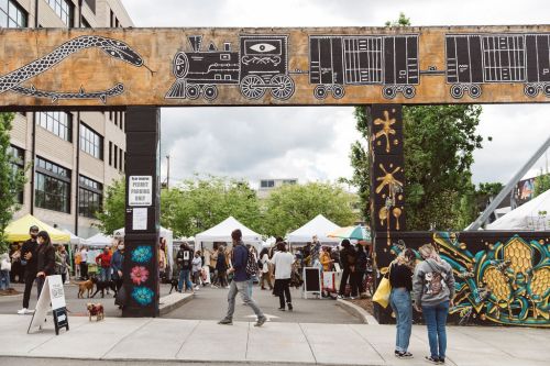 A vibrant outdoor market scene with people walking under a colorful archway, surrounded by tents and street art.