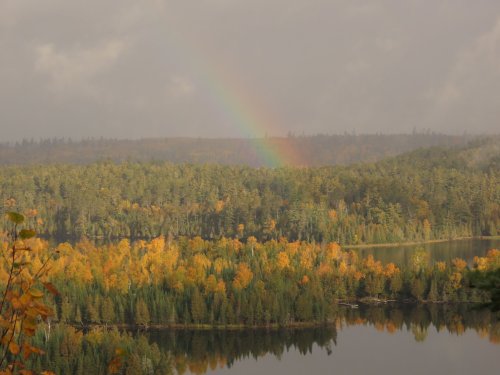A serene landscape featuring a rainbow over a forest of autumn-colored trees and a calm lake.
