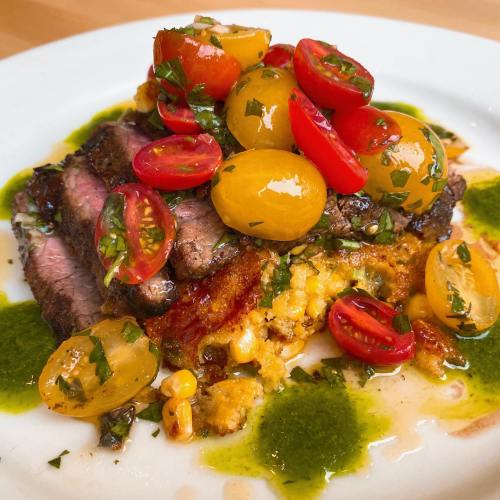 A plate of grilled steak topped with cherry tomatoes and herbs, served over a bed of corn and drizzled with green sauce.
