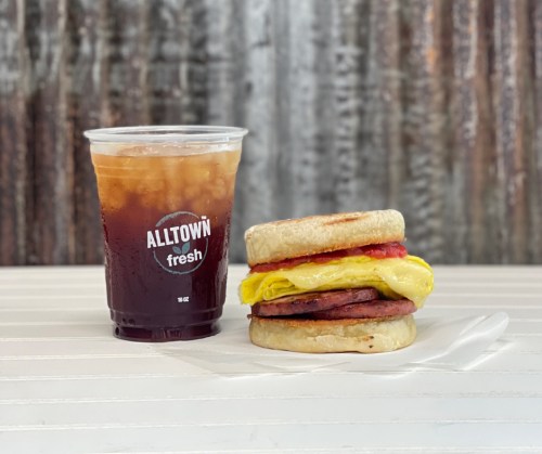 A breakfast sandwich with egg, ham, and tomato next to a cup of iced coffee on a white table.