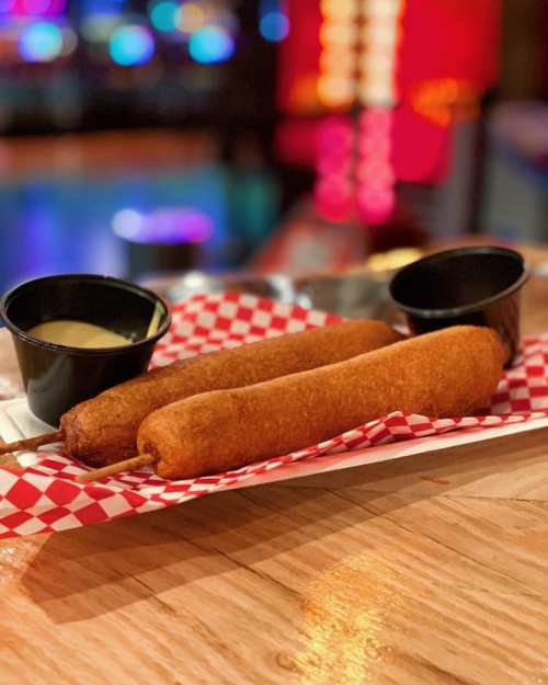 Two golden corn dogs on a paper tray, accompanied by two small cups of dipping sauce. Colorful lights in the background.
