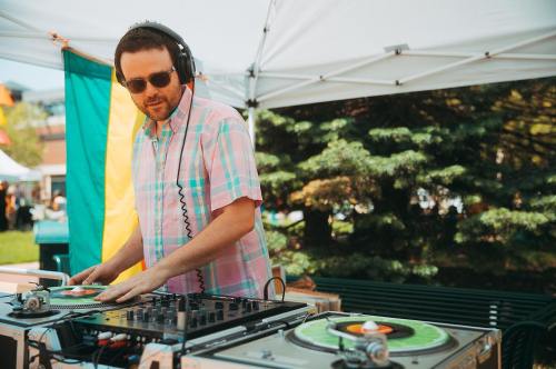 A DJ in sunglasses mixes music at an outdoor event, wearing a colorful plaid shirt under a tent.