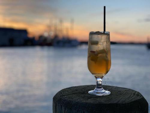 A refreshing cocktail in a glass sits on a wooden post, with a sunset and boats in the background.
