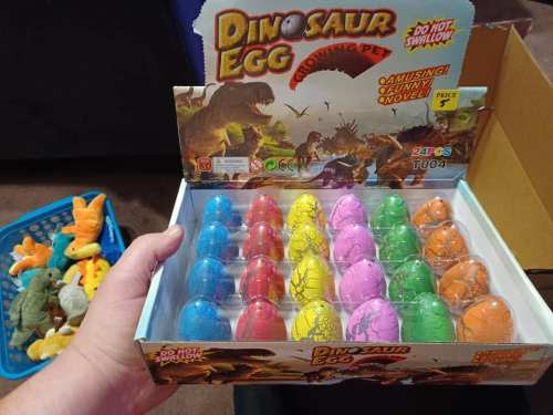 A hand holds a box of colorful dinosaur egg toys, with a basket of plush dinosaurs in the background.