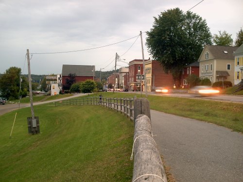 A quiet street scene with a grassy area, wooden fence, and charming buildings under a cloudy sky.