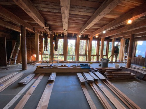 Interior of a construction site with wooden beams, planks on the floor, and large windows showing a forest view.
