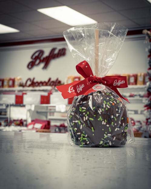 A chocolate-covered apple wrapped in clear plastic and tied with a red ribbon, displayed in a candy shop.