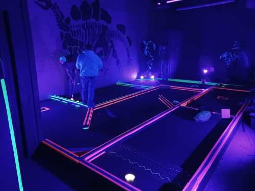 A glow-in-the-dark mini-golf course with neon lines and a dinosaur mural, featuring players in a darkened room.