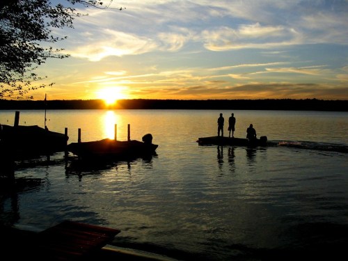 Silhouettes of people by a lake at sunset, with boats and a colorful sky reflecting on the water.