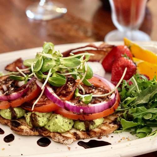A colorful plate featuring avocado toast topped with tomatoes, onions, sprouts, and a side of fresh fruit.