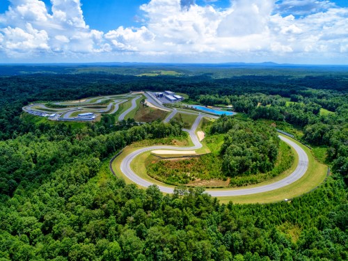 Aerial view of a winding racetrack surrounded by lush green forests and blue skies with scattered clouds.