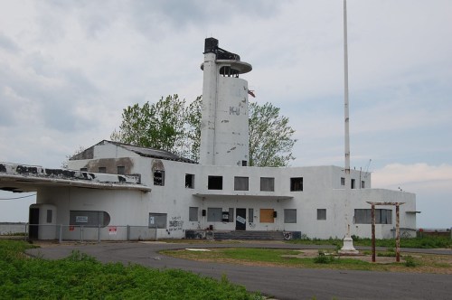 Abandoned white building with a tower, broken windows, and overgrown greenery, set against a cloudy sky.