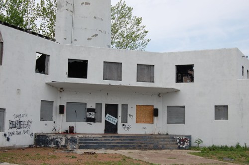 Abandoned building with boarded windows, graffiti, and a staircase leading to a closed entrance. Overcast sky above.