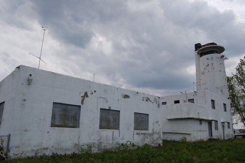 Abandoned white building with a tower, surrounded by overgrown grass and under a cloudy sky.