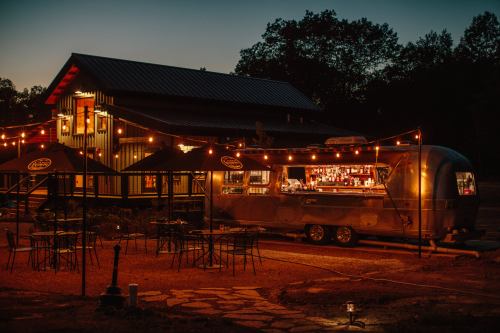 A vintage Airstream bar with string lights, surrounded by outdoor seating, near a modern building at dusk.