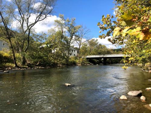 A serene river scene with trees, a bridge, and a house in the background under a partly cloudy sky.
